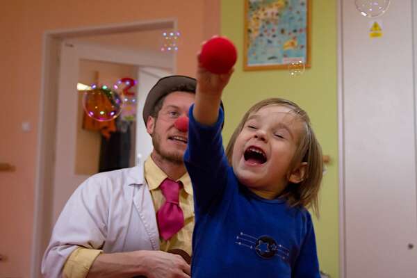 A small boy holds a red nose in his hand and is catching the bubbles in the hospital romm as a clown is joyfully watching him.