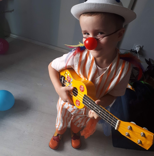 Little Leo wears a red nose, a hat and plays the ukulele in his clown costume.