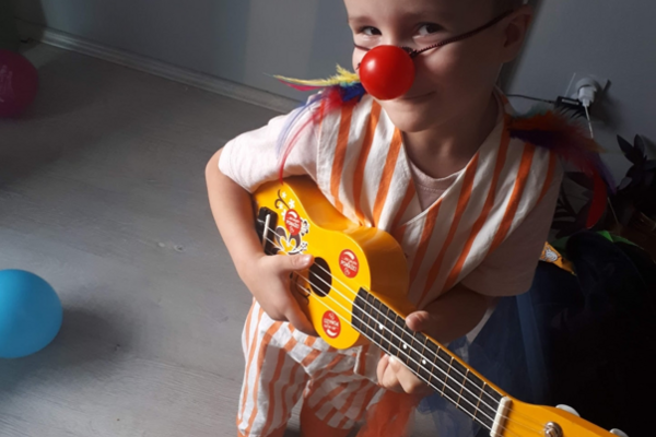 Little Leo wears a red nose, a hat and plays the ukulele in his clown costume.