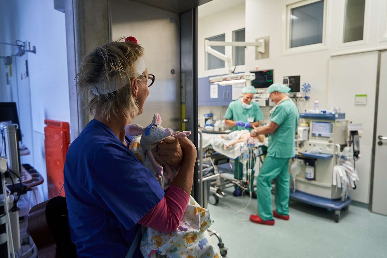 A female RED NOSES clown holds a tiny material animal in her had and watches the doctors prepare a little boy for surgery
