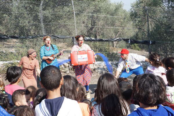 RED NOSES clowns are in the middle of a show and a large audience is watching them as they make music and spread a blue scarf on the stage