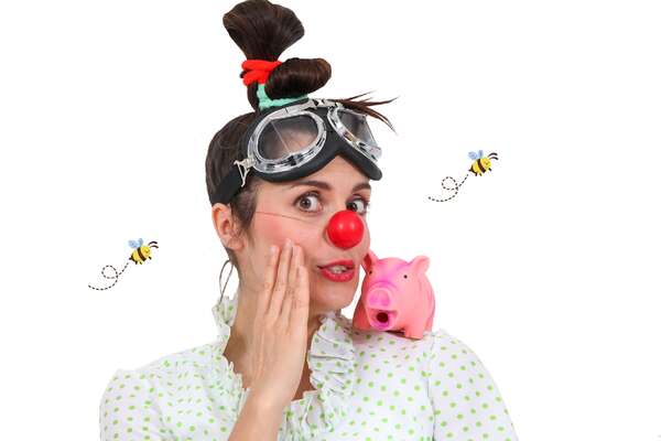 A female clown with a red nose touches her face as she looks into the camera and has a small pink piglet on her shoulder
