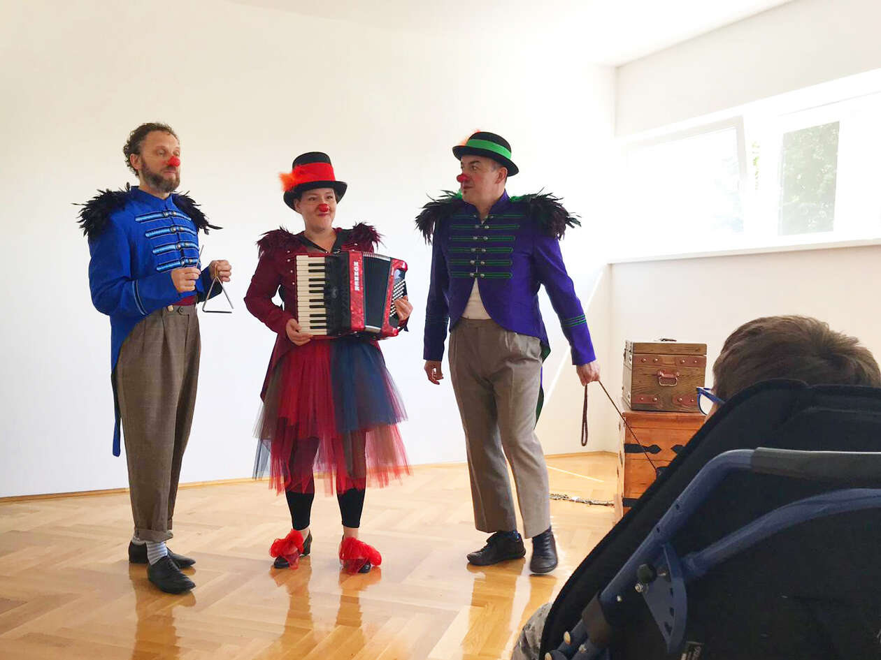 Three RED NOSES clowns perform a song in front of the children present in the room.