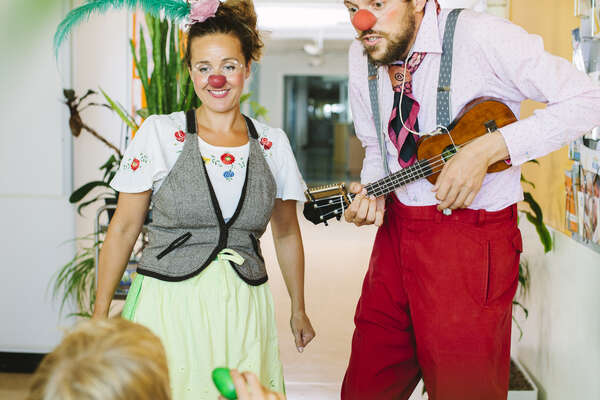 A boy with a purple T-Shirt hold a green egg in his hand while two RED NOSES clowns make music with the ukulele and look joyfully at the boy