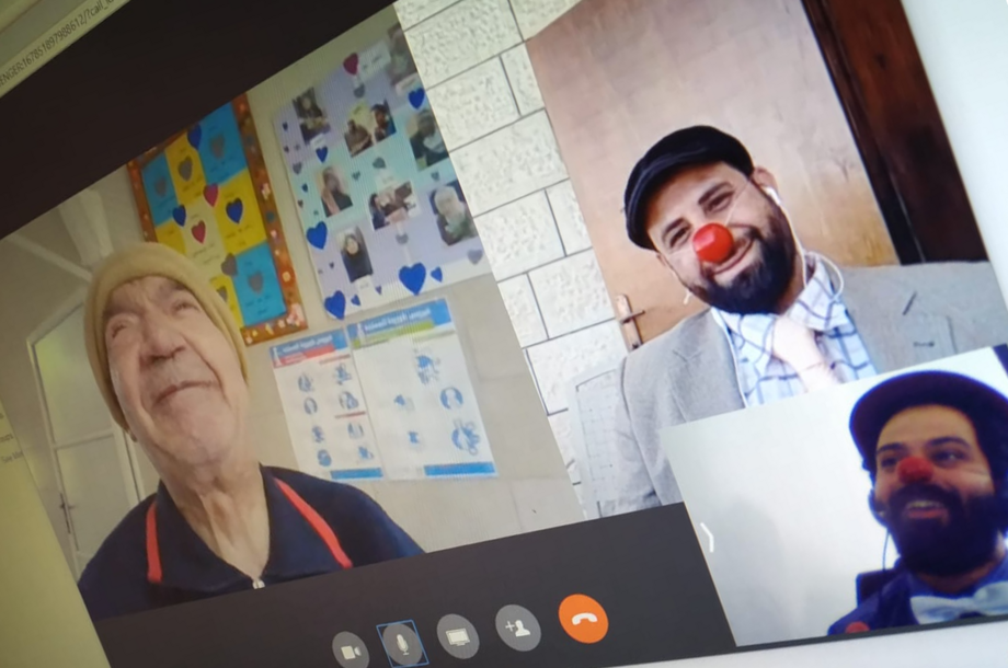 Two male clowns from RED NOSES Palestine smile during an online clowning visit to an elderly man whose face is bright and joyful.
