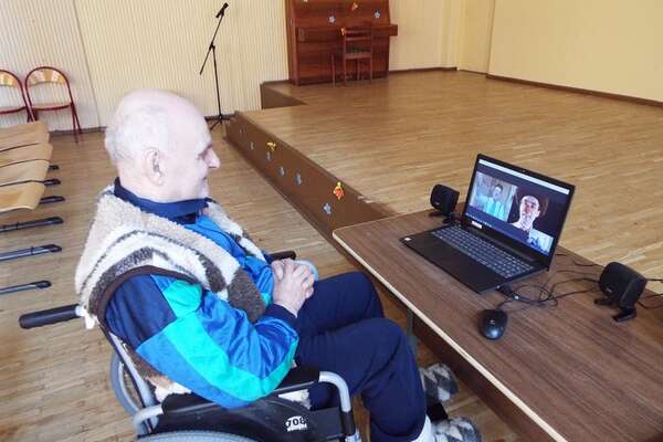 An old man in an elderly care home sits in front of the laptop in a big empty room and watches a clown from RED NOSES Lithuania perform a show online 