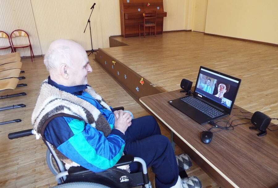 An old man in an elderly care home sits in front of the laptop in a big empty room and watches a clown from RED NOSES Lithuania perform a show online 