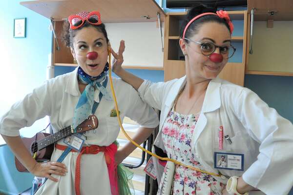 Two female clowns stand together in a room and one is touching the ear of the other in a playful way as if it were testing her hearing ability.