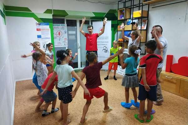 Children are standing in a circle in a big room and are learning tricks from the clowns (spinning plates).