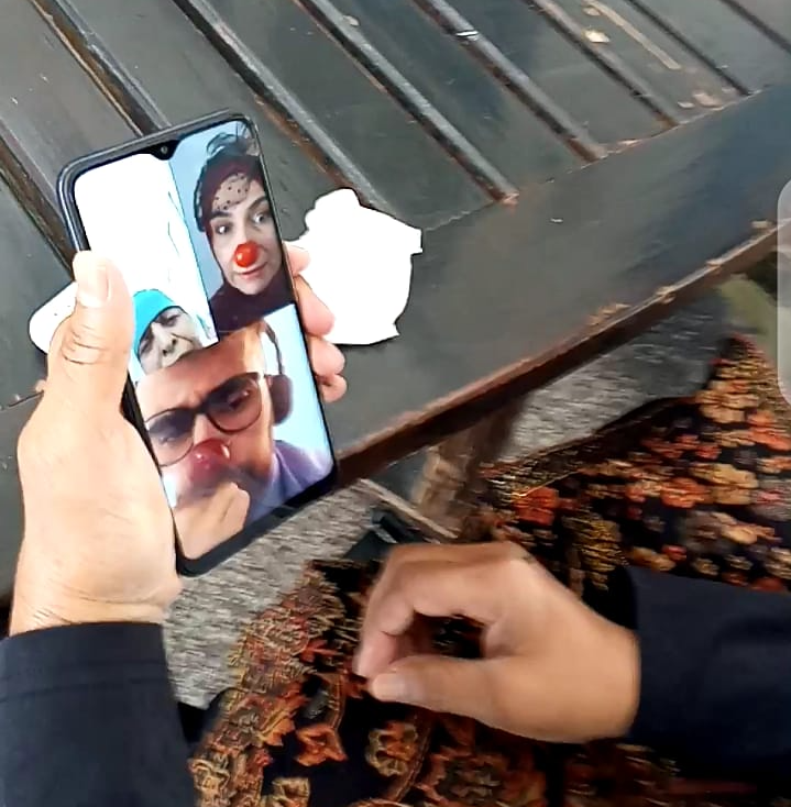 An older woman in Palestine watches two clowns on the screen of a mobile phone and smiles.