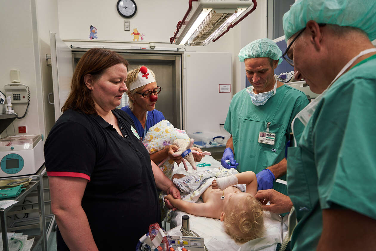 Doctors, the mother and the RED NOSES female clown stand around the bed where little Luca is prepared for surgery.
