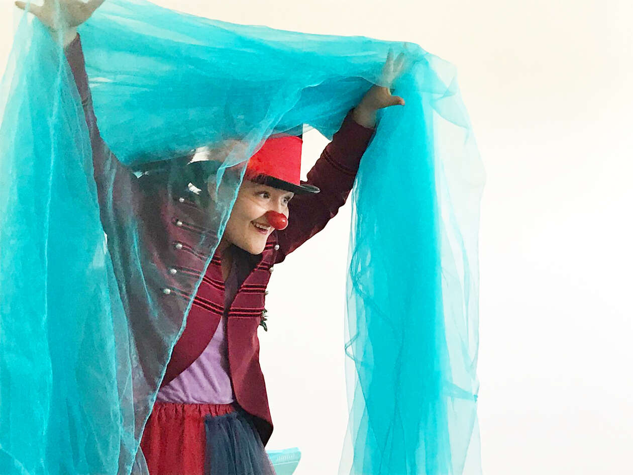 A female clown with a big red hat and a red nose holds a blue, light silk curtain with her hands.