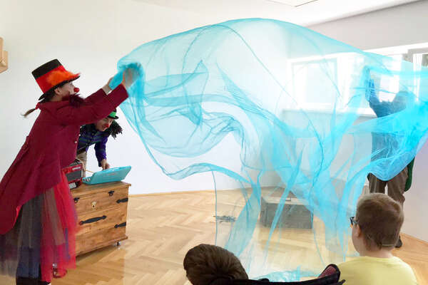Two RED NOSES clowns are spreading a blue silk curtain in a big room, being watched by the children there.