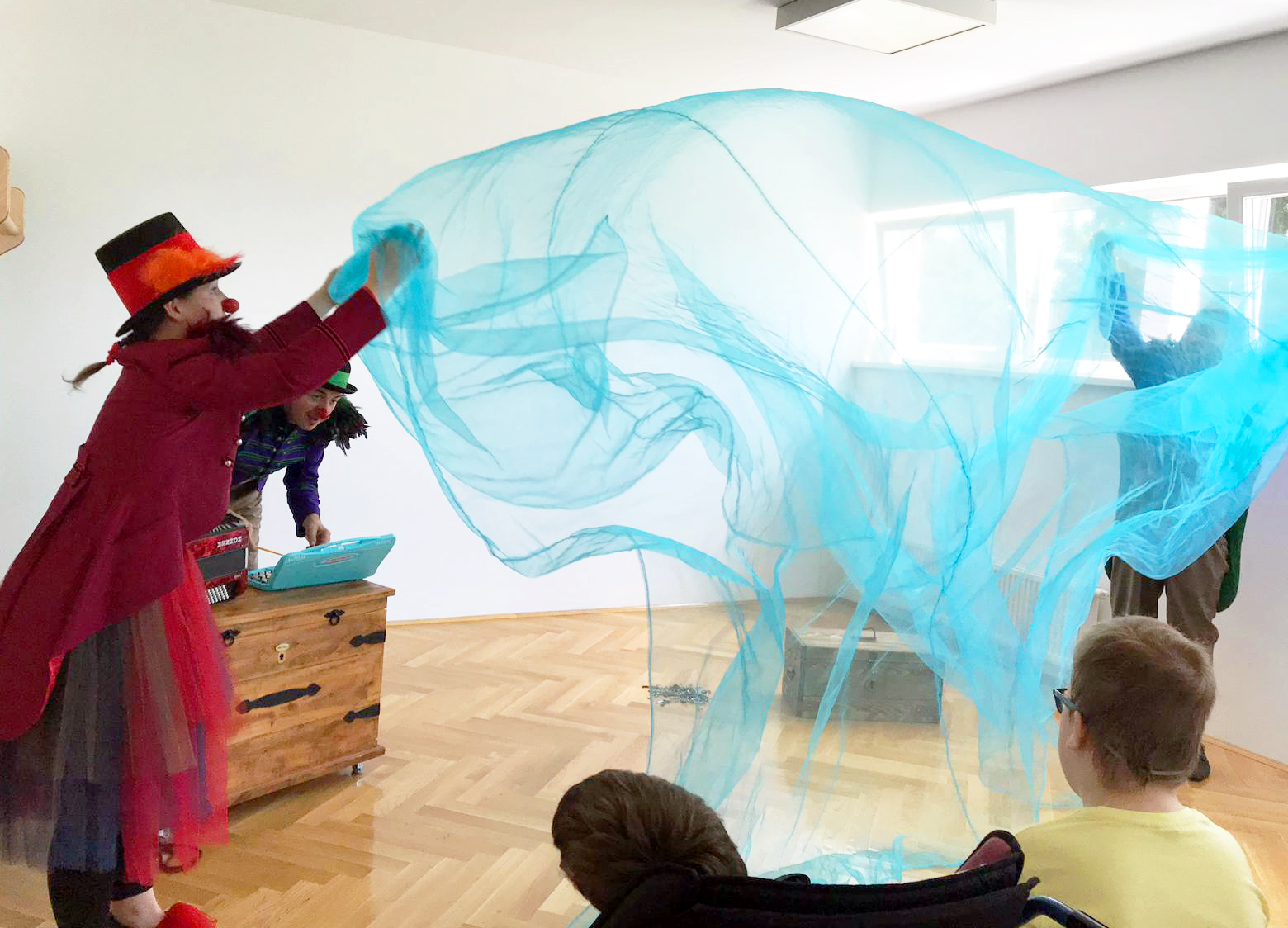Two RED NOSES clowns are spreading a blue silk curtain in a big room, being watched by the children there.