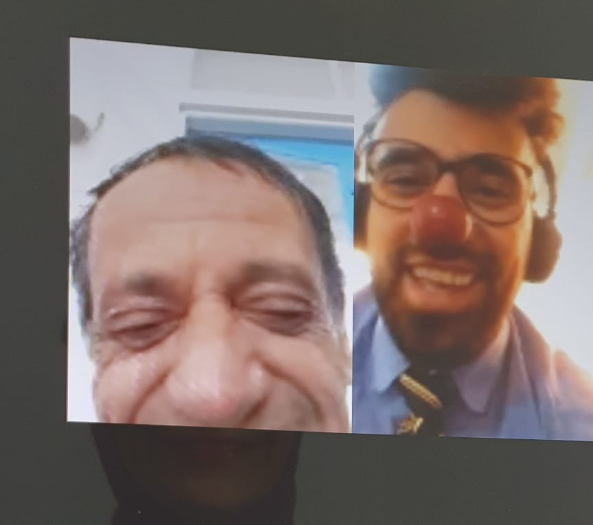 An older man in an elderly home in Palestine is encountered joyfully by a male clown and smiles.